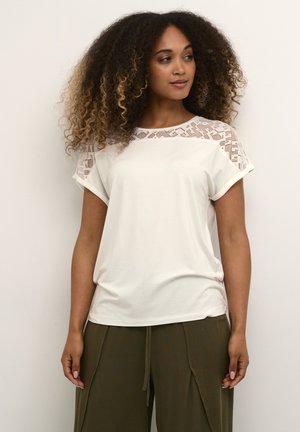 Short-sleeve cream T-shirt with lace detailing on shoulders, smooth fabric, relaxed fit, paired with loose olive green trousers.