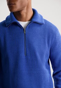 Pulover albastru cu fermoar, cu textură ribbed și guler înalt. Are un închidere cu fermoar la jumătate și un aspect de țesătură moale.