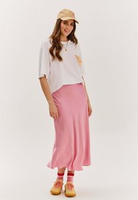 Bílý zkrácený tričko s oranžovou grafikou, růžová saténová maxi sukně, pruhované růžové a červené ponožky a žluté tenisky. Béžová čepice s logem.