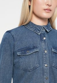 Denim krekls gaiši zilā krāsā, ar pogu rindu, norādītu apkakli un divām krūšu kabatām. Raksturo gluda tekstūra ar nelielām izbalēšanas pazīmēm.