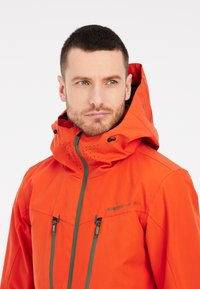 Giacca impermeabile arancione con colletto alto e cappuccio regolabile. Presenta tasche con zip e perforazioni traspiranti sul cappuccio.