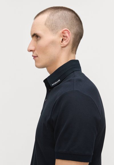 Marinblå pikétröja med krage, korta ärmar och en märkesaccent på kragen. Tillverkad av texturerat tyg och har en knappstängning framtill.