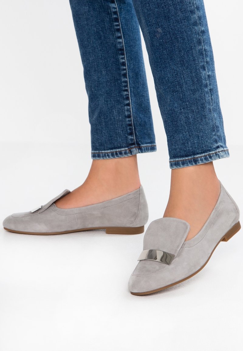 Des pieds portant des mocassins en daim gris avec des détails de boucles métalliques, associés à un jean en denim bleu, se tenant sur une surface blanche.