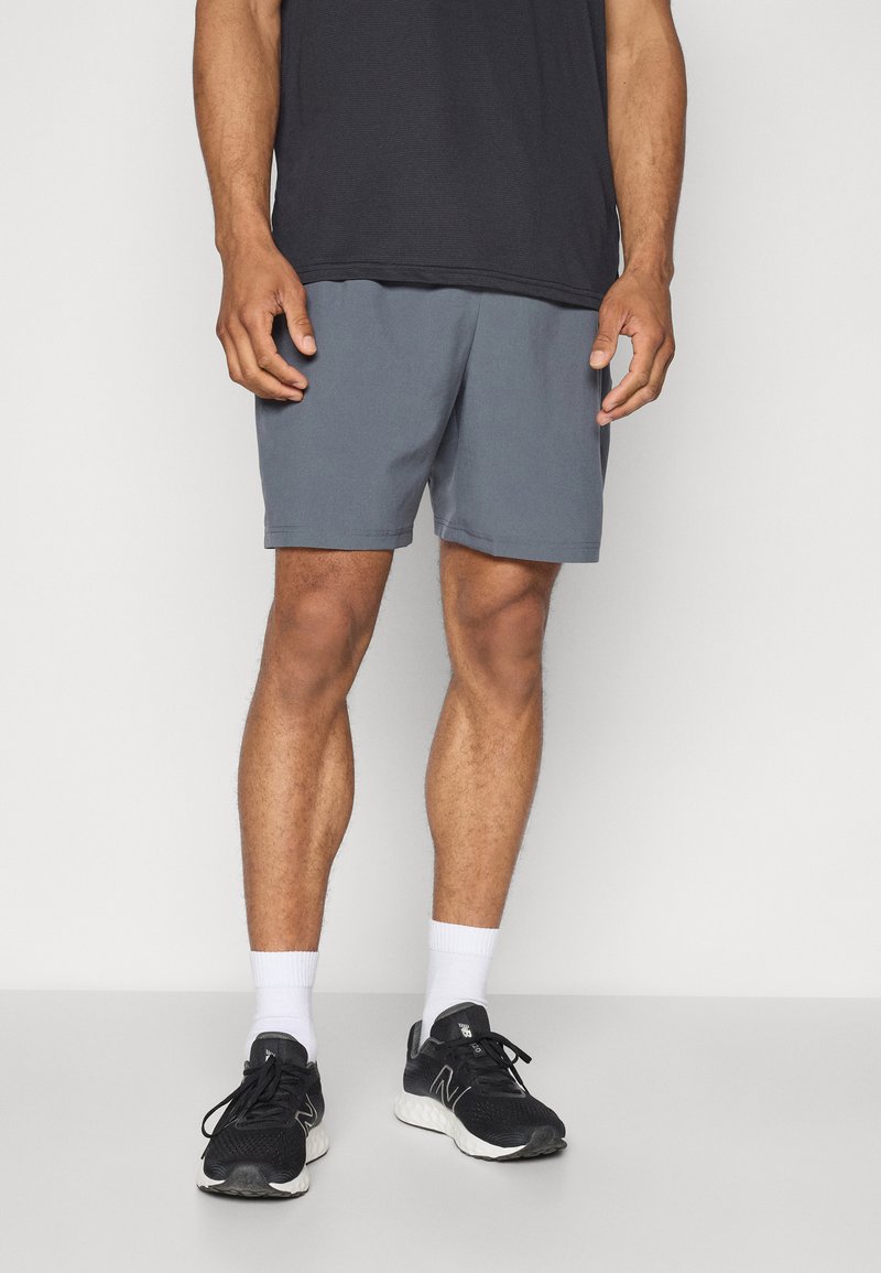 Man som bär grå shorts, svarta sneakers, vita strumpor och en svart t-shirt, stående mot en enkel vit bakgrund.