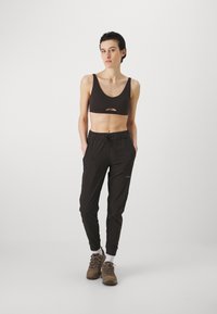 Melnā sporta krūšturis ar priekšējo izgriezumu, kombinēts ar brīvā stila melnām sporta biksēm ar elastīgu jostu un sānu kabatām. Brūni pārgājienu zābaki.