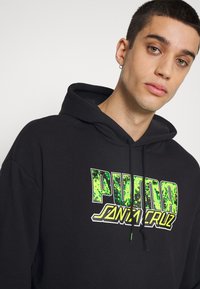 Felpa nera con cappuccio caratterizzata da una grande stampa del logo verde e bianco che riporta la scritta "PUMA SANTA CRUZ" sul davanti. Materiale in cotone morbido.