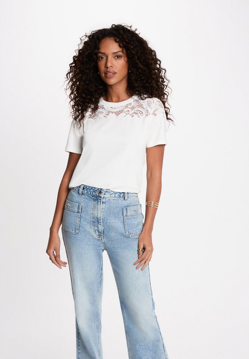 Woman with curly hair wearing a white short-sleeve top with lace detail and light blue high-waisted jeans with front patch pockets.
