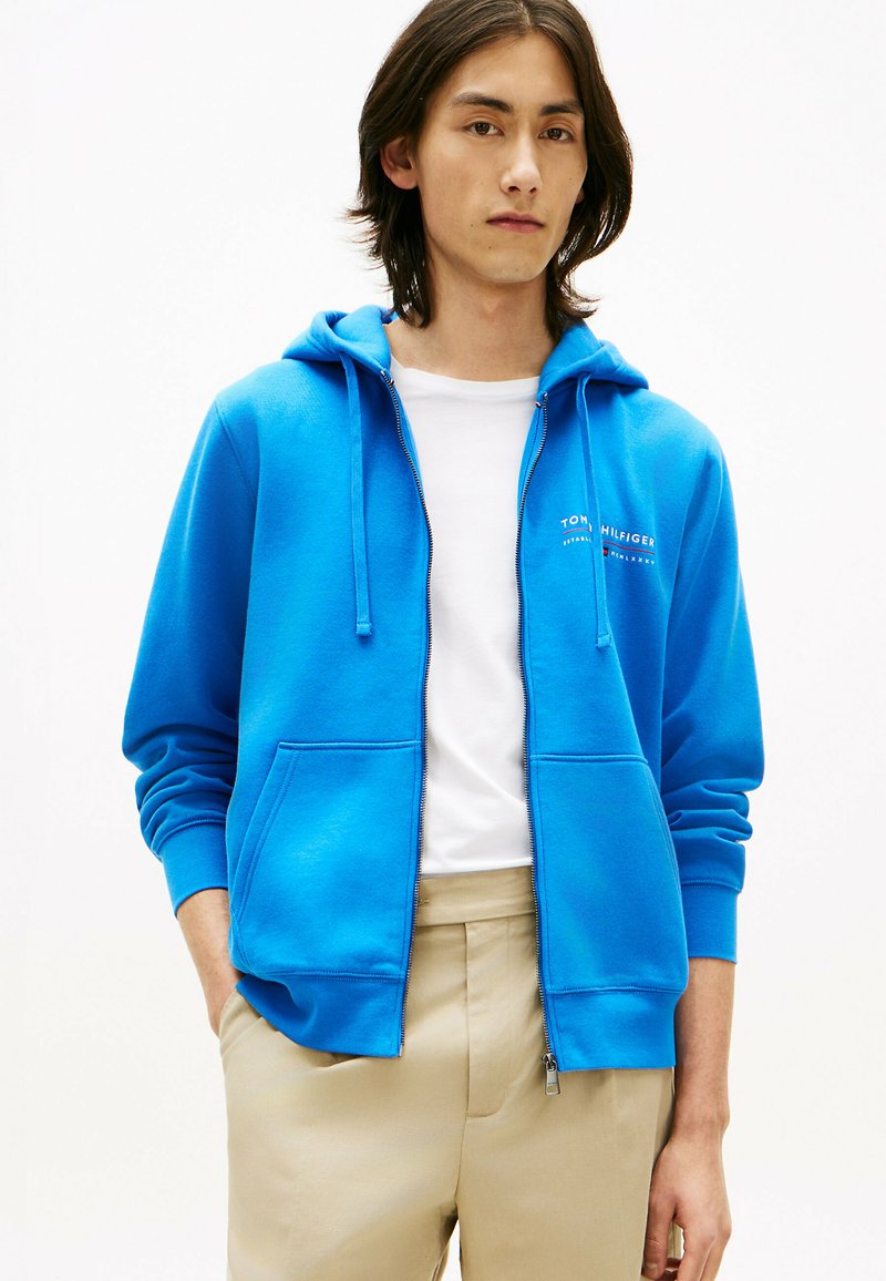 Jeune personne portant un sweat à capuche zippé bleu vif par-dessus un t-shirt blanc et un pantalon beige, debout avec les mains dans les poches devant un fond uni.
