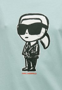 Κινούμενο σχέδιο ενός ατόμου με μεγάλα γυαλιά ηλίου, μαλλιά χτενισμένα προς τα πίσω και κοστούμι, που στέκεται με τα χέρια στις τσέπες πάνω από το κείμενο "KARL LAGERFELD".