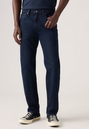 Mand iført mørkeblå jeans og sorte lave sneakers står mod en ensfarvet baggrund.
