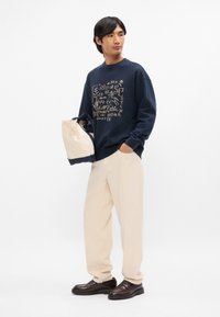 Ναυτικό φούτερ με χρυσό γραφικό σχέδιο, κρέμα παντελόνι και σκούρα καφέ παπούτσια. Κρατάει μία κρέμα και ναυτική τσάντα. Απλή, άνετη εφαρμογή.