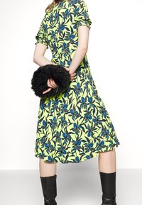 Donna in un abito midi floreale giallo tiene una borsa nera pelosa dietro la schiena, indossando stivali neri fino al ginocchio su uno sfondo bianco.