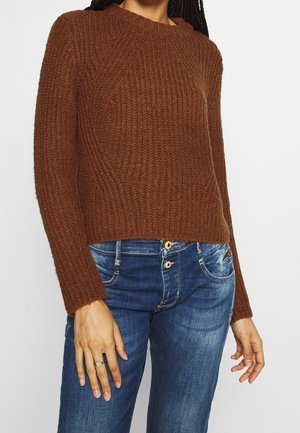 Personne portant un pull en maille côtelée marron et un jean en denim bleu, avec une bague en or à la main gauche.