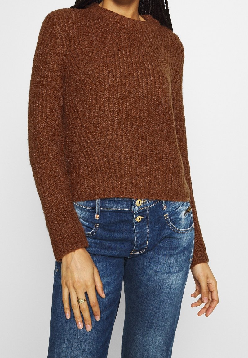 Personne portant un pull en maille côtelée marron et un jean en denim bleu, avec une bague en or à la main gauche.