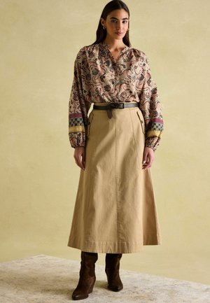 Langärmelige, gemusterte Bluse in Creme und Braun, kombiniert mit einem beigen Midikleid. Die braunen Wildleder-Stiefel mit Absatz runden das Outfit ab.