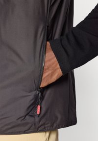 Chaqueta negra con una textura suave y ligera y un bolsillo lateral con cremallera; cuenta con una manga oscura en contraste y un detalle de tirador de cremallera rojo.