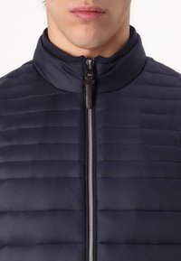 Μπλέ φουσκωτό μπουφάν με οριζόντιες ραφές, ψηλό γιακά και μπροστινό φερμουάρ. Το ύφασμα φαίνεται απαλό και ελαφρύ.