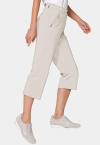 Pantalons crops beige clair avec une poche latérale, devant plat et coutures surpiquées, associés à des chaussures de sport gris clair. Texture de tissu lisse.