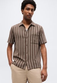 Άνδρας που φοράει καφέ πουκάμισο με κοντά μανίκια και λευκές κάθετες ρίγες, και μπεζ παντελόνι, στέκεται με τα χέρια στις τσέπες.