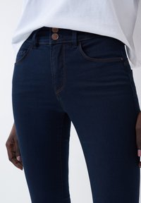 Jean skinny bleu foncé en tissu mélangé coton, avec des coutures orange, deux boutons à la taille et des poches avant fonctionnelles.