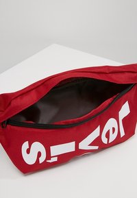 Pochette con zip rossa realizzata in tessuto resistente, caratterizzata da grandi lettere bianche su un lato e una fodera interna nera liscia.