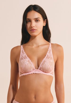 Mujer con un bralette de encaje floral rosa con tirantes finos sobre un fondo beige.