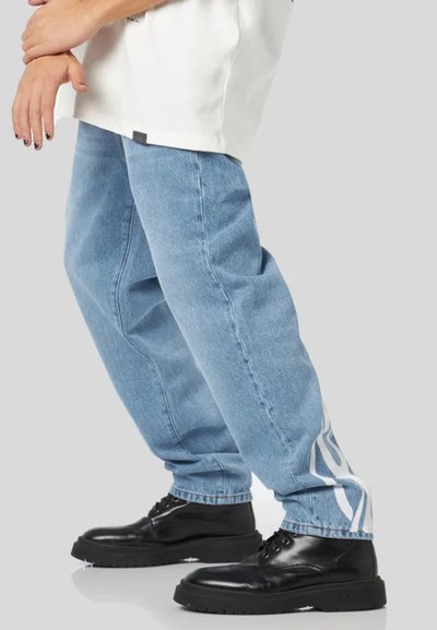 Jeans in denim blu chiaro con una vestibilità ampia, caratterizzati da un motivo a fiamma bianca di lato, abbinati a stivali neri con lacci.