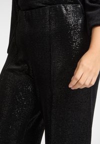 Czarne spodnie z cekinami o teksturowanej powierzchni, z prostym krojem i widoczną kieszenią. Ręka spoczywa na tkaninie, podkreślając jej połysk.