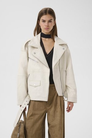 Naine kannab ülisuurte mõõtmetega valget jalgratturi jopet, musta V-kaelusega topi kaelarihma detailiga, pruune tekstuuriga pükse, hoides käes sobivat käekotti.