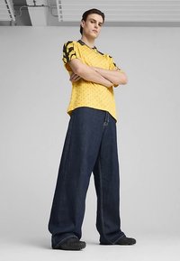 Žlutá košile s krátkými rukávy s geometrickým vzorem a černými akcenty, nošená s nadměrnými tmavě modrými džínami a texturovanými černými botami.