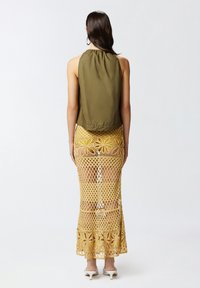 Maslina zelena bez rukava majica u kombinaciji s dugom žutom čipkastom suknjom s cvjetnim uzorcima. Model nosi bijele sandale na petu.