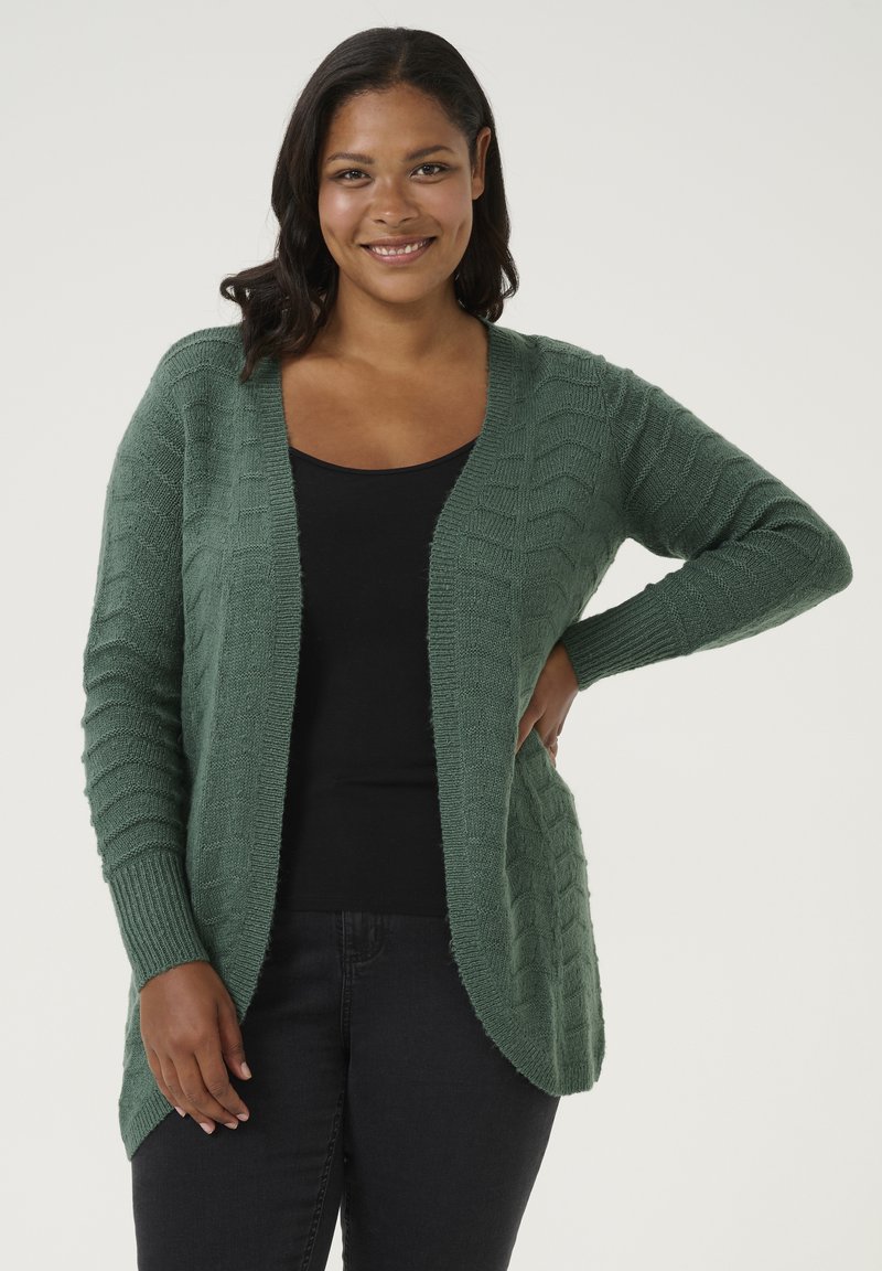 Gebreide cardigan in donkergroen met een textuurpatroon, open voorkant, lange mouwen en geribde manchetten, gelegd over een zwarte top.