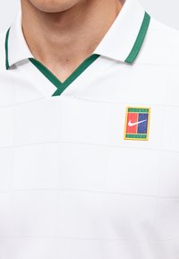 Homme portant une chemise blanche à col avec une bordure verte et un logo Nike rectangulaire coloré sur la poitrine gauche.
