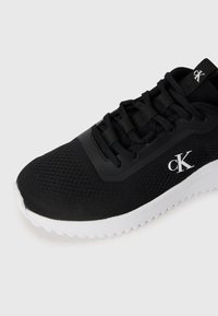 Juodos Calvin Klein sportbačiai su tinklo viršumi, juodomis raištelėmis ir balta padu, rodantys pirštų ir šonų logotipą šviesiame fone.