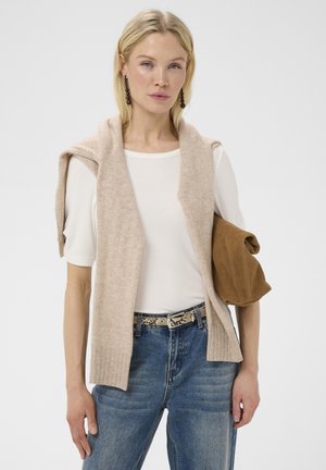 Lyshåret kvinde iført hvid kortærmet top, beige strikket vest over skuldrene, blå jeans, mønstret bælte, holder en brun sammenfoldet jakke på armen.