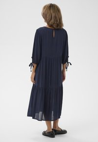 Marineblaues, gestuftes, knielanges Kleid mit Dreiviertelärmeln, einem Schlüsselrücken-Detail und Bindebändern an den Ärmelabschlüssen. Tragen mit schwarzen Schuhen.