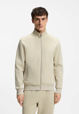 Ung man klädd i en beige zip-up-jacka och matchande byxor, stående med ett neutralt uttryck mot en enkel vit bakgrund.