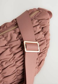 Gevoerde roze schoudertas met een textuurafwerking, voorzien van een gouden ritssluiting en een brede stoffen schouderband met een rechthoekige metalen gesp.