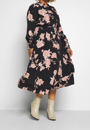 Vrouw draagt een zwarte midikleding met roze bloemenprint, lange mouwen en een tailleband, gecombineerd met lichtgekleurde enkellaarzen.
