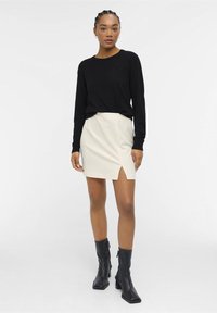 Sort langærmet top parret med en lys beige mini-nederdel med en sideslids. Modellen bærer sorte hæle støvler. Glat stoftekstur.