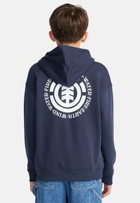 Niño con sudadera azul marino con un logo blanco circular y las palabras "Agua Fuego Tierra Viento" en la espalda, combinado con pantalones cortos de mezclilla azules.