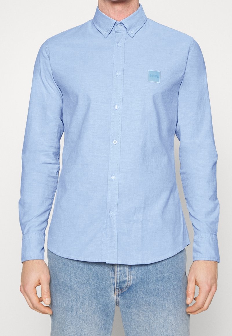 Valkoinen nappi-yläosa, jossa on pitkät hihat, terävä kaulus ja pieni neliön muotoinen logo vasemmassa rinnassa. Puuvillamateriaali, sileä rakenne.