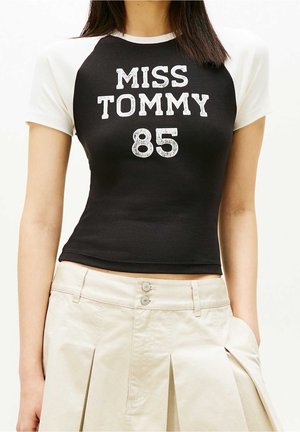 Kobieta w czarno-białej bluzce z krótkim rękawem z napisem "MISS TOMMY 85" i beżowych plisowanych spodniach z wysokim stanem, z ręką w kieszeni.