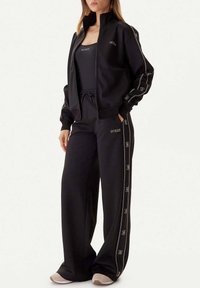 Zwart trainingspak met een jas met ritssluiting en hoge kraag en broek met zijstrepen. Het materiaal is zacht, met geborduurde logodetails.