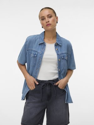 Vrouw staat met handen in de zakken, draagt een lichtblauw denim kortemouwenhemd, een wit hemdje en zwarte denimshorts tegen een witte achtergrond.