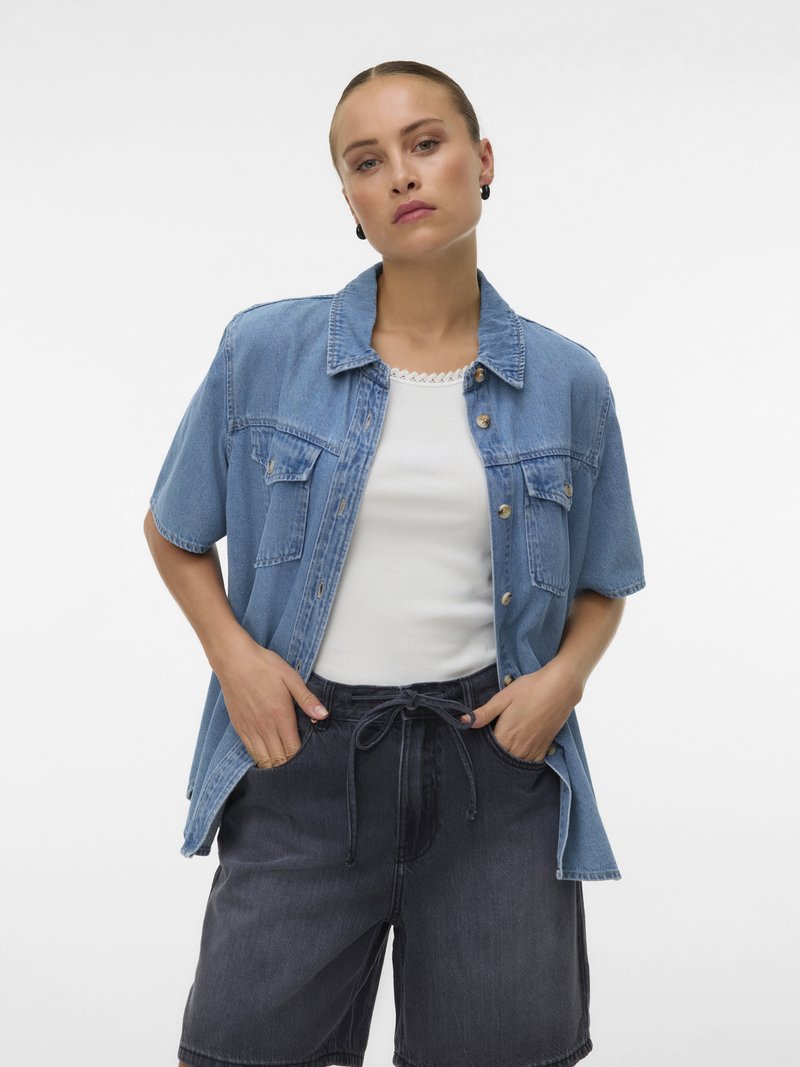 Naine seisab käed taskutes, seljas helesinine lühikeste varrukatega teksasärk, valge tanktop ja mustad teksapüksid valgel taustal.