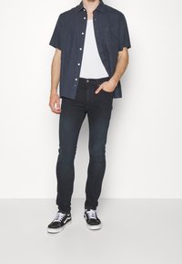 Camisa de botón de manga corta azul oscuro sobre una camiseta blanca, combinada con jeans ajustados oscuros y zapatillas negras y blancas con cordones.