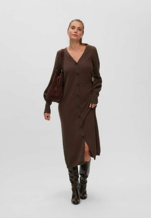 Femme portant une longue robe marron boutonnée, tenant un sac à main marron assorti et des bottes hautes noires, debout devant un fond uni.