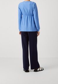 Modrý texturovaný dlouhý rukáv s nařaseným pasem, spárovaný s tmavomodrými širokými kalhotami a černými sandály, pohled zezadu.