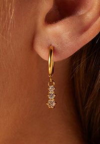 Gouden creool met een hangende ketting met drie sprankelende heldere kristallen, met een gladde textuur en een gepolijste afwerking.